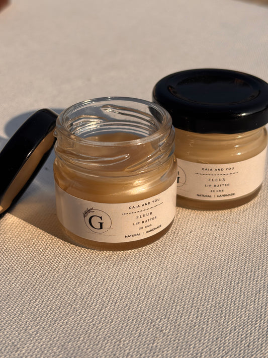 Two small glass jars with black lids, labeled 'Fleur Lip Butter', one jar is open showing the solid lip butter inside, the other is closed.