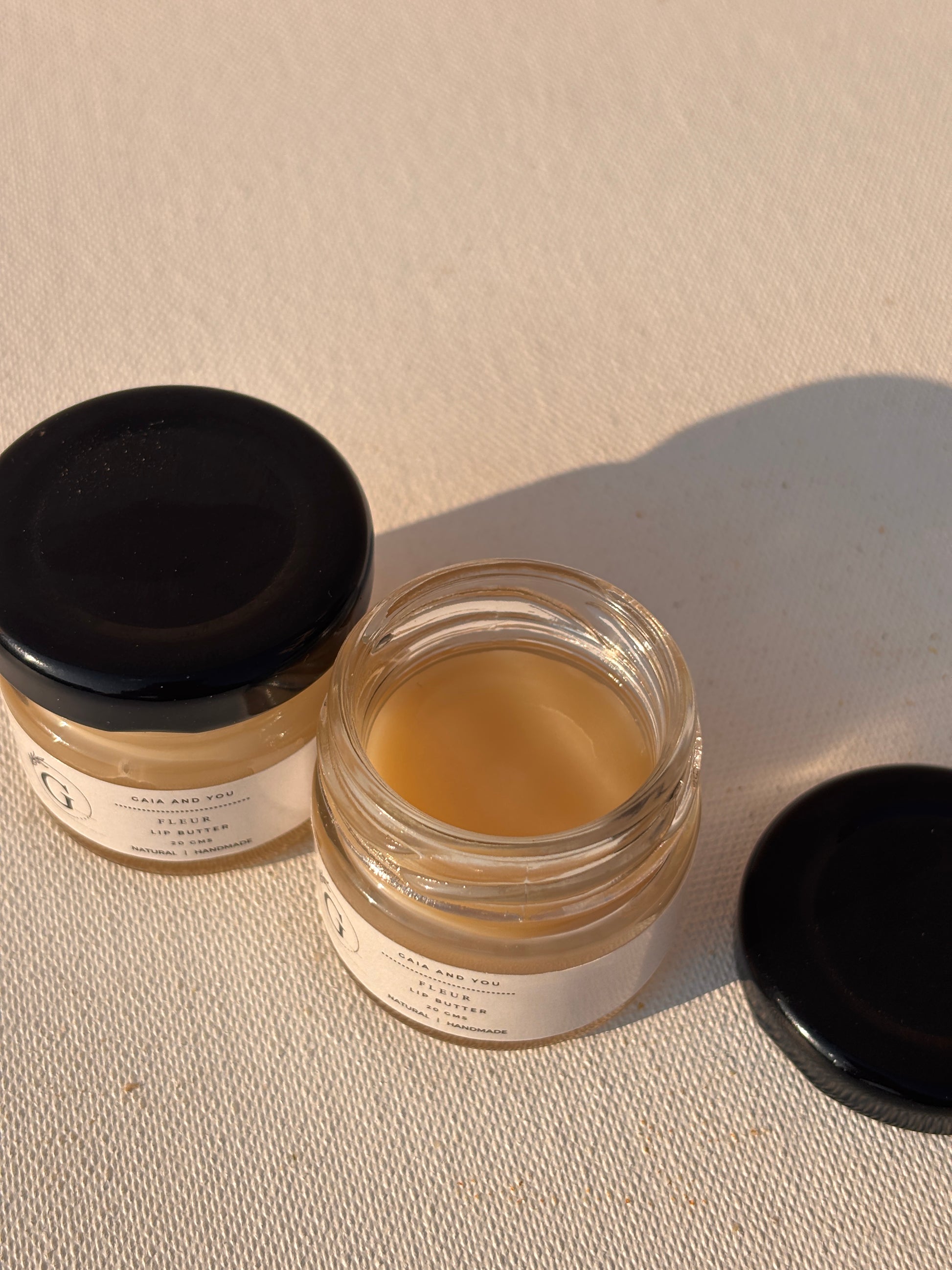 Two small glass jars with black lids, labeled 'Fleur Lip Butter', one jar is open showing the solid lip butter inside, the other is closed.