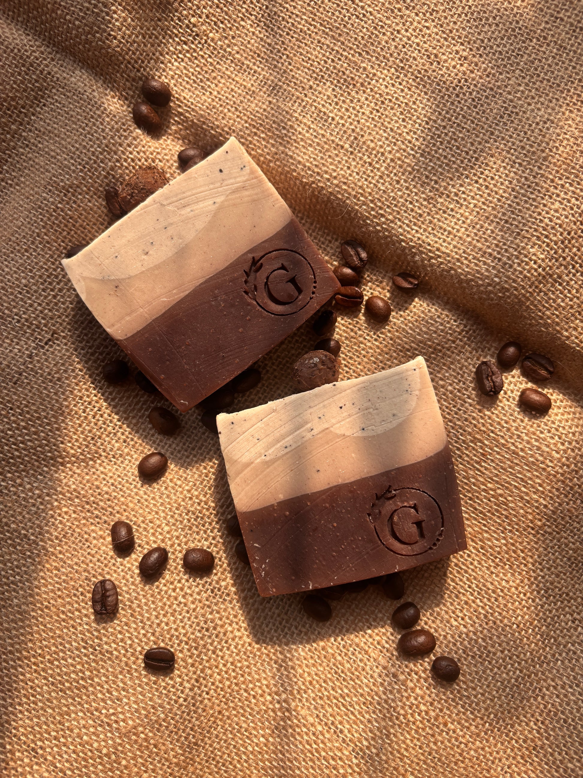 Two artisanal coffee soaps made with finest Chikmaglur coffee on a burlap with scattered coffee beans. The soaps are of gradient earthy brown going from darker to lighter because of the intense coffee decoction and moisturising kokum butter.