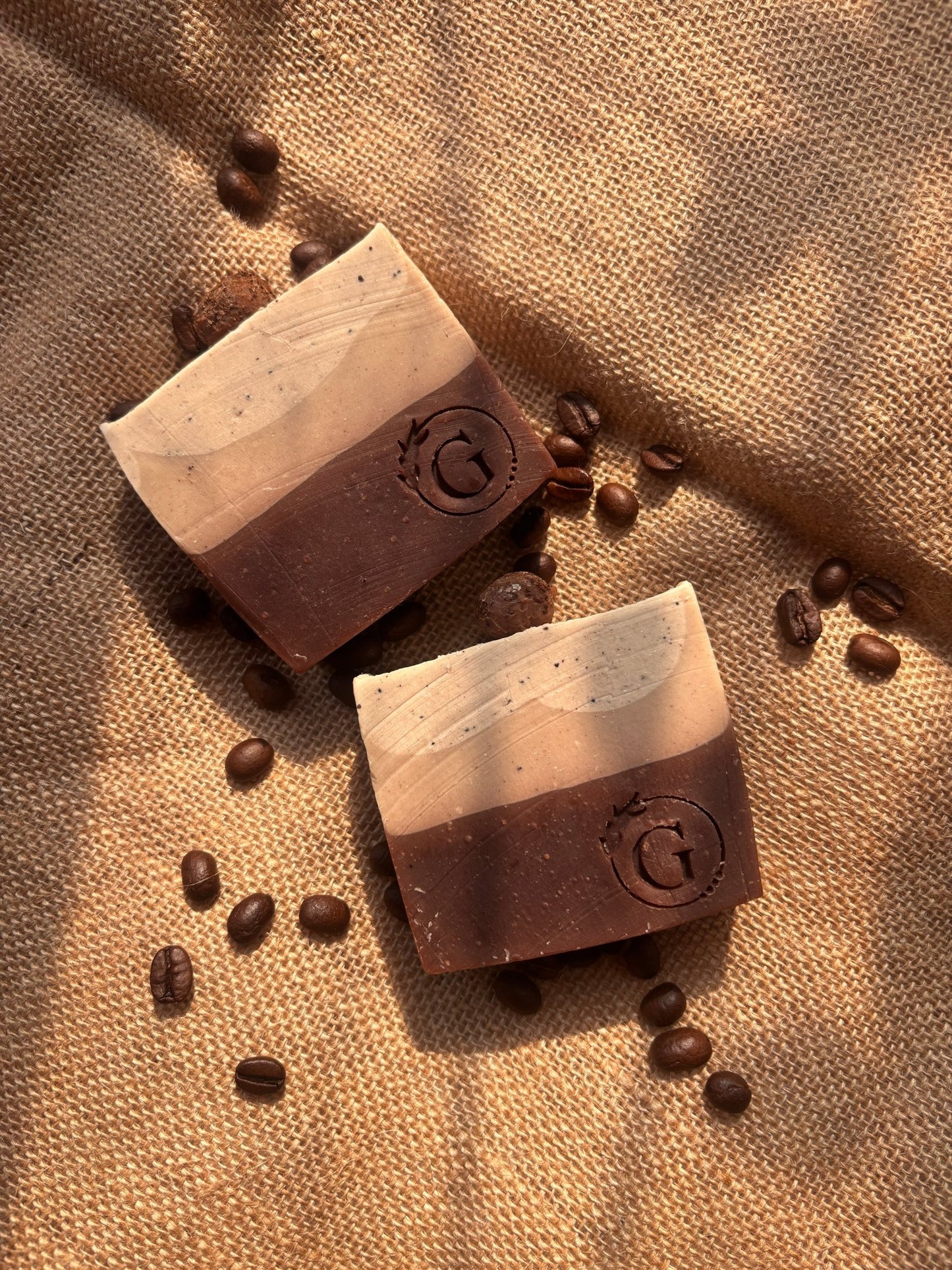 Two artisanal coffee soaps made with finest Chikmaglur coffee on a burlap with scattered coffee beans. The soaps are of gradient earthy brown going from darker to lighter because of the intense coffee decoction and moisturising kokum butter.